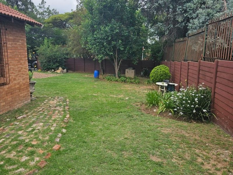 A private rear garden featuring a brick-walled apartment exterior, paved walkway, established trees, and perimeter timber fencing.