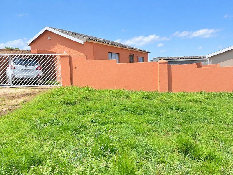 Front view of a single-story house featuring a perimeter wall, metal gate, and a tiled roof, set behind an unmaintained grass lawn.