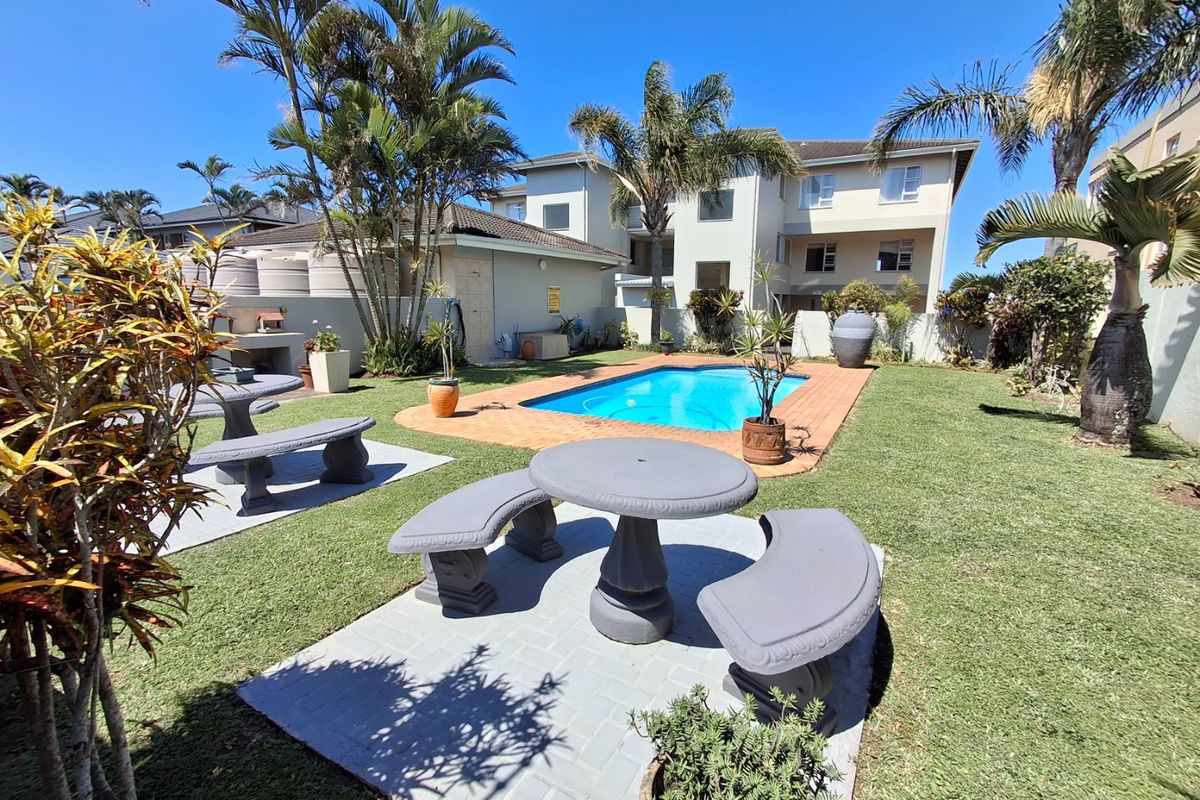 A well-maintained communal pool area featuring brick paving, a built-in braai, and established garden landscaping within an apartment complex setting.