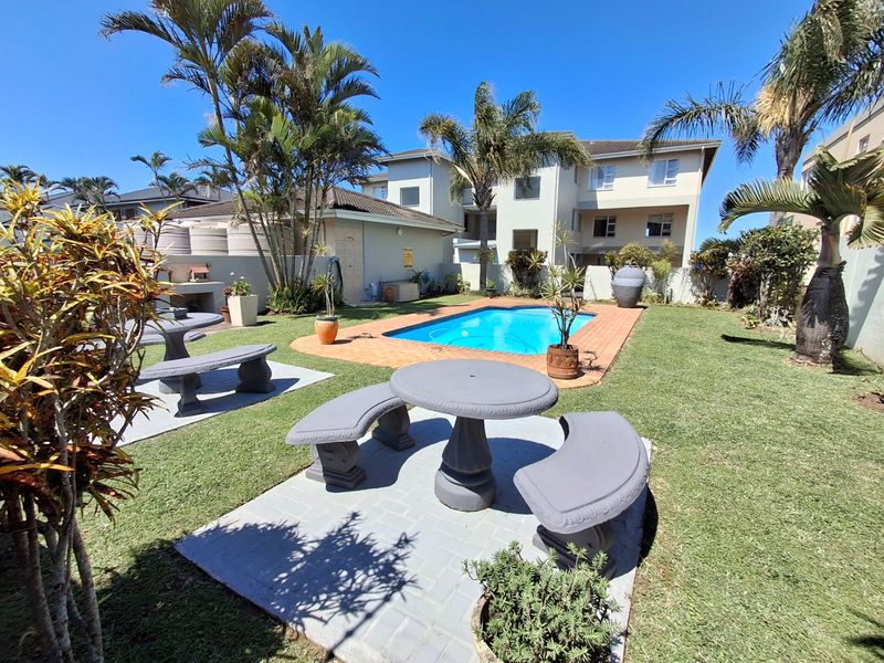 A well-maintained communal pool area featuring brick paving, a built-in braai, and established garden landscaping within an apartment complex setting.