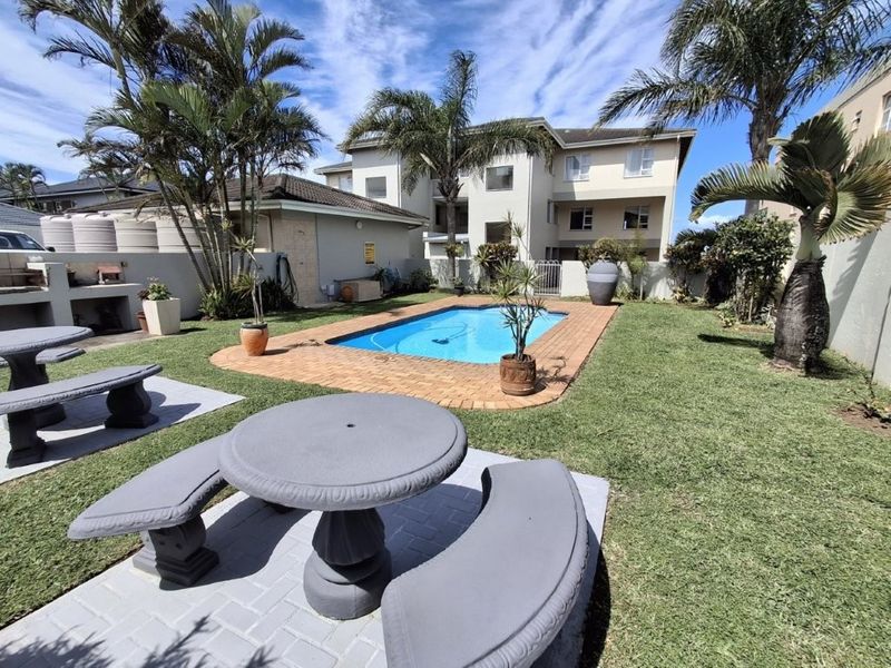 Outdoor communal area featuring a swimming pool, paved patio, built-in braai, and established garden landscaping within an apartment complex.