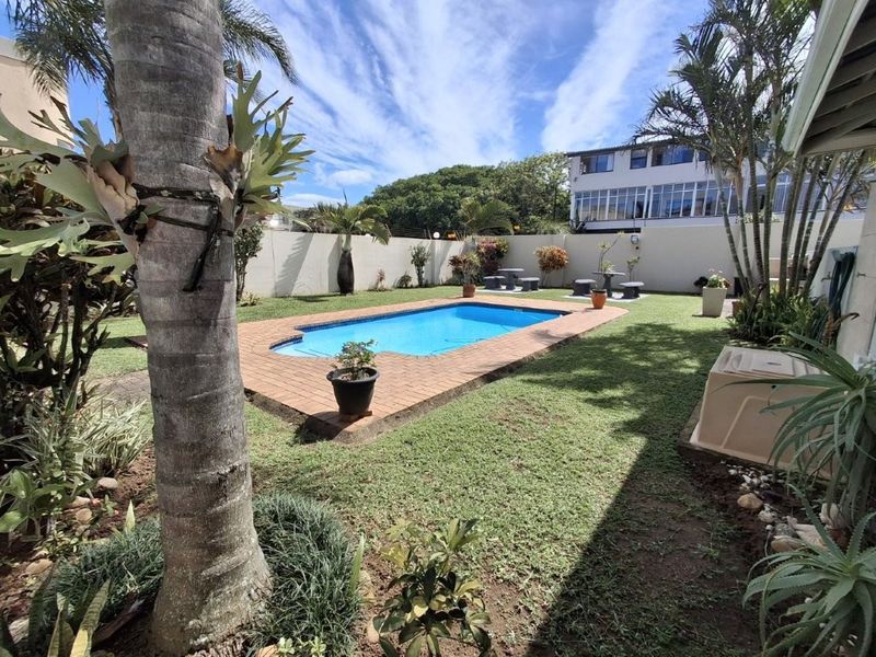 A well-maintained outdoor pool area featuring a swimming pool with brick coping, surrounded by a lawn and perimeter wall.