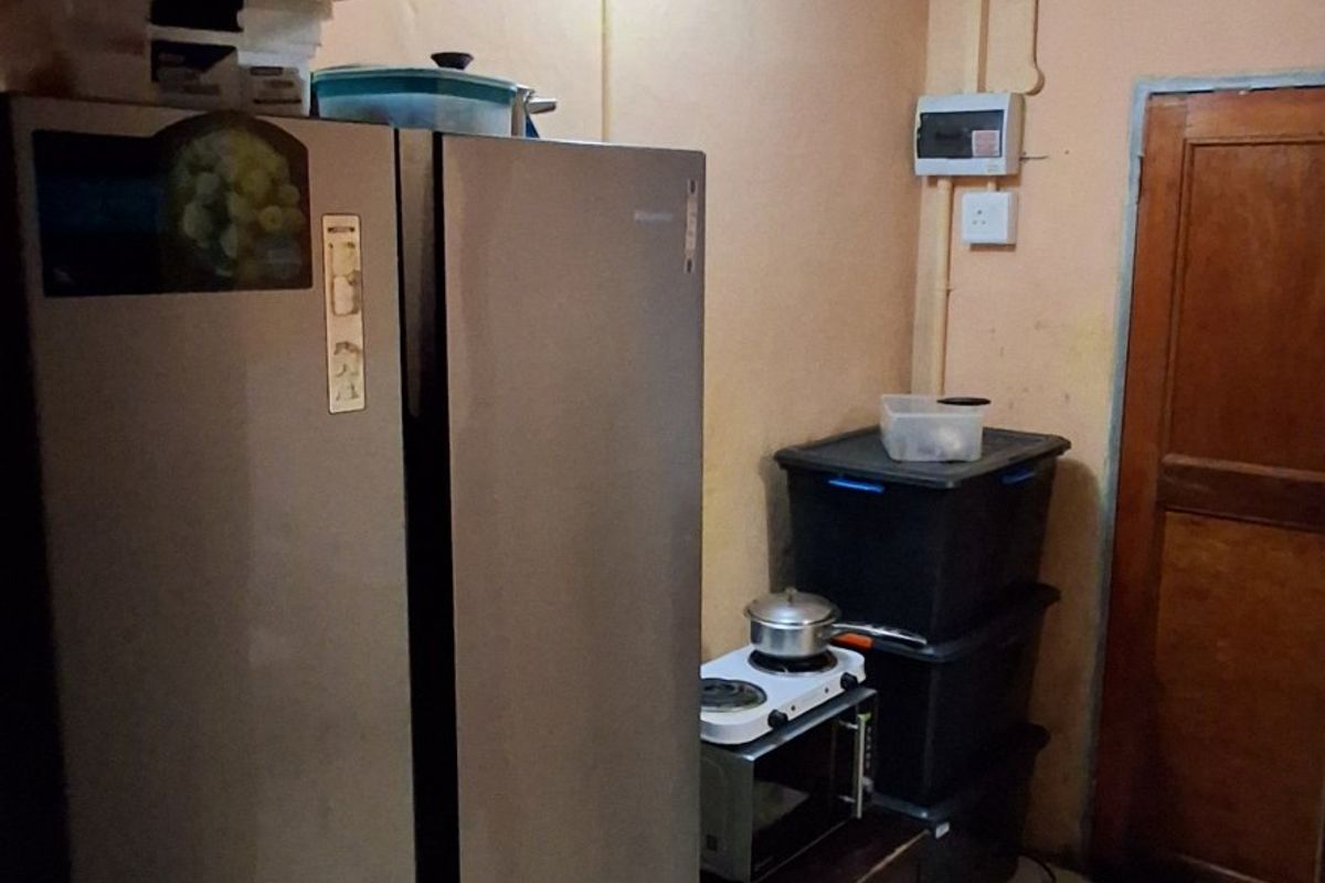 A small kitchen area featuring tiled flooring, a wooden door, and an exposed electrical distribution board, requiring cosmetic updates and improved lighting.