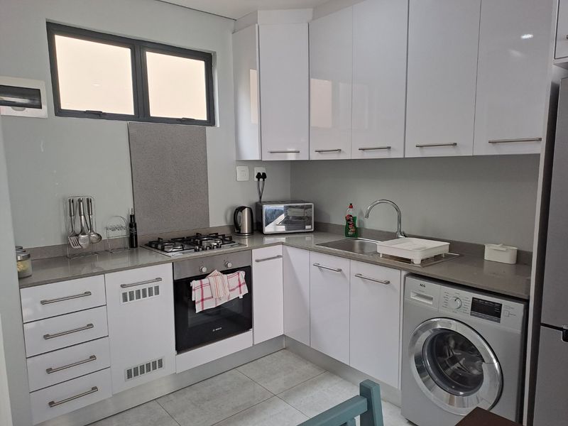 A modern, compact kitchen featuring white cabinetry, stone countertops, a gas hob, built-in oven, and integrated laundry appliance space.