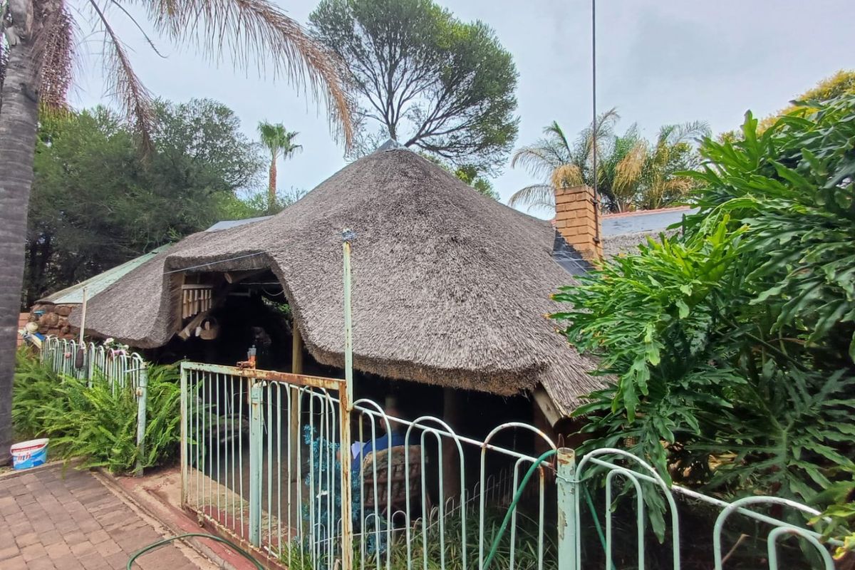 Rear view of the property featuring a traditional thatch roof structure, brick chimney, and surrounding garden area with metal perimeter fencing.