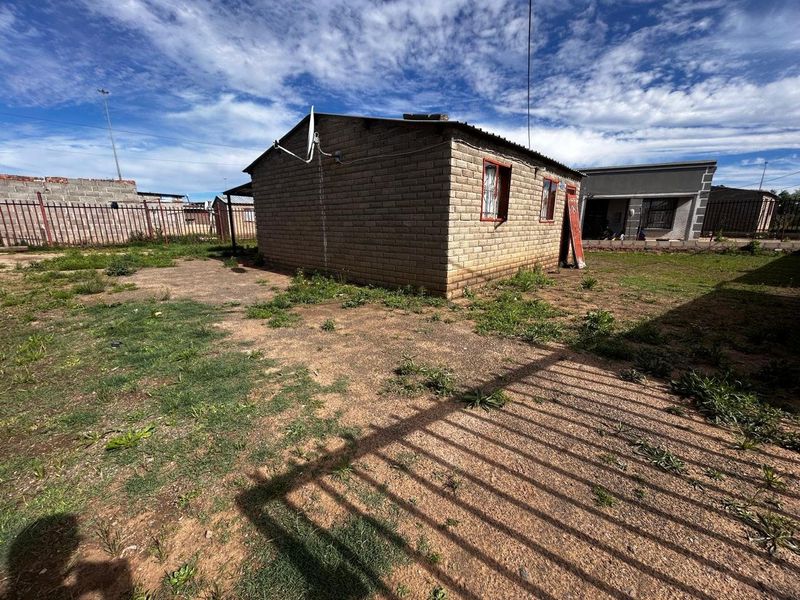 Rear view of a detached brick house featuring a corrugated metal roof, satellite dish, and surrounding metal perimeter fencing on a large plot.