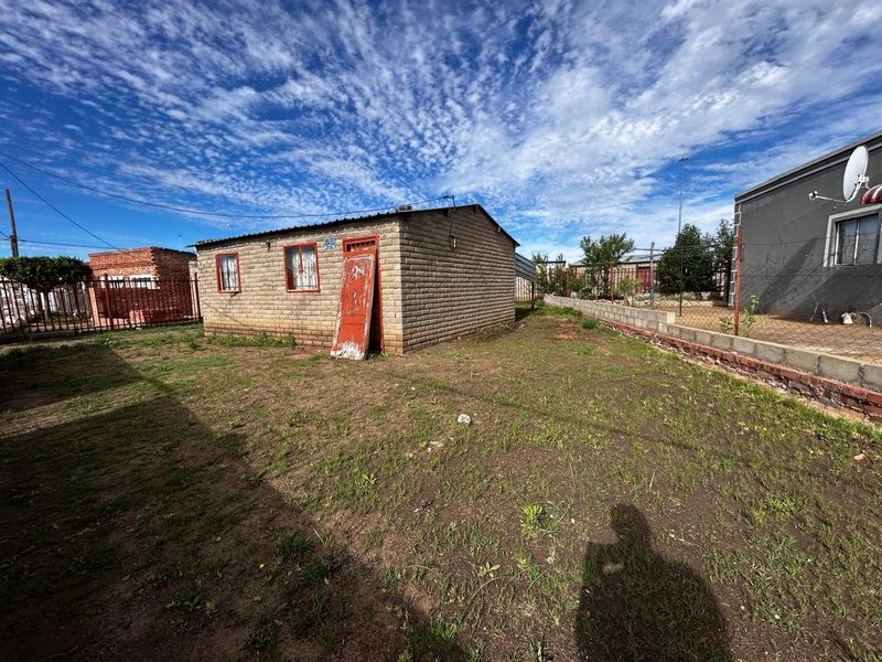 Rear view of a detached brick structure with a corrugated metal roof, set on a large plot with wire fencing and boundary walls.