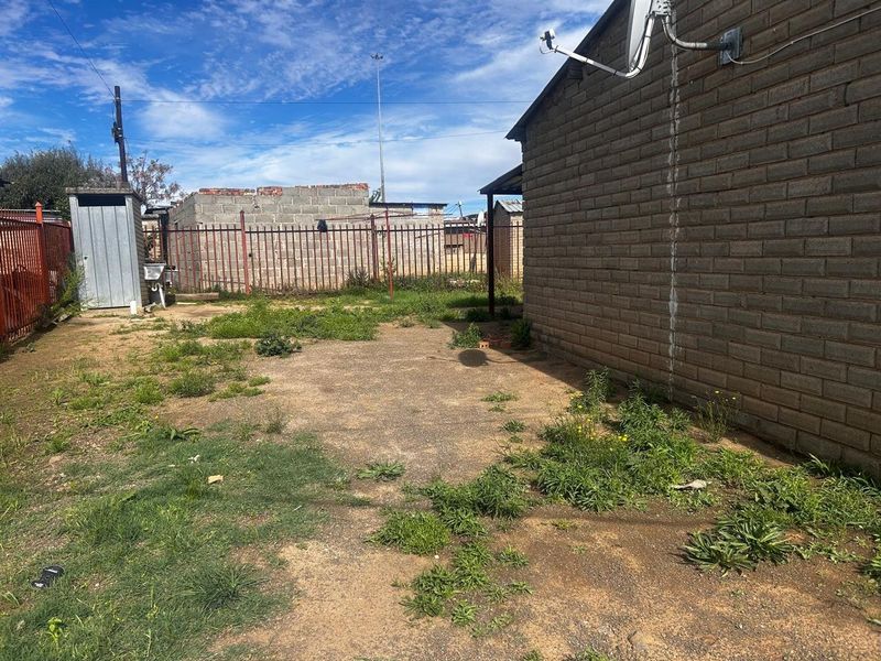 A rear exterior view of the property featuring a brick facade, metal perimeter fencing, and an outdoor utility shed in a large yard area.
