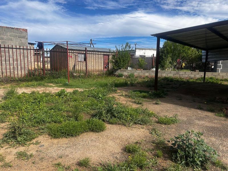A spacious rear yard featuring a metal carport, perimeter steel fencing, and an outbuilding, offering significant potential for landscaping and outdoor improvement.