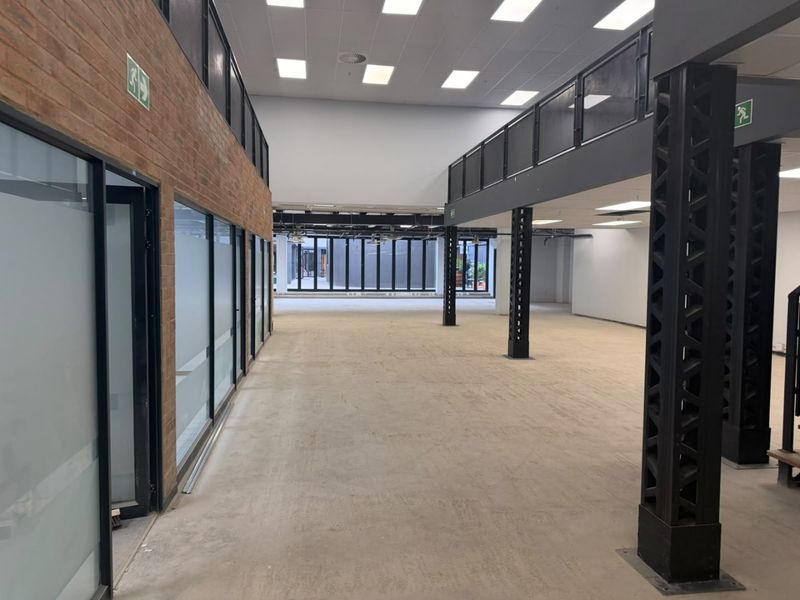 Spacious open-plan commercial office area featuring industrial design elements, exposed brickwork, steel columns, and a mezzanine level with glass partitions.