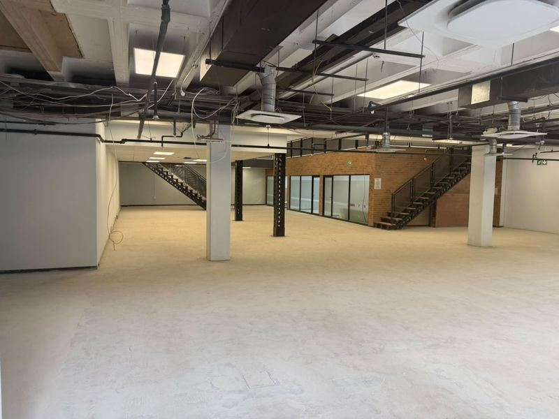 A large, open-plan commercial space featuring exposed industrial ceilings, structural columns, two staircases, and glass-partitioned office areas, currently in shell condition.