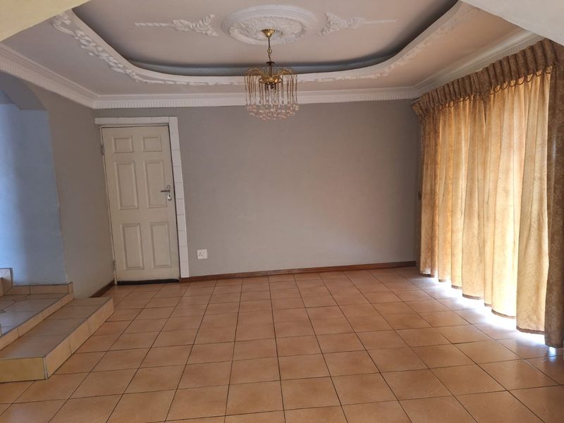 Spacious living area featuring tiled flooring, ornate ceiling detailing with a central light fixture, and access to an adjacent space via a doorway.