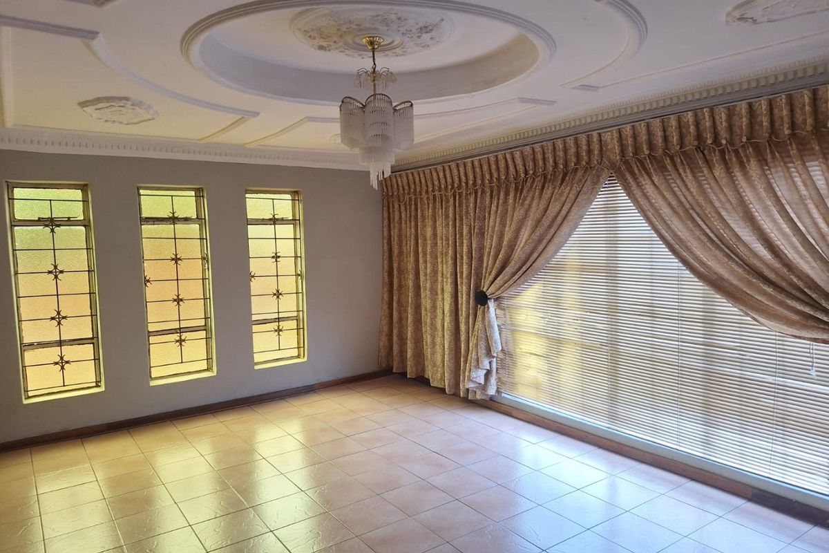 Spacious living room featuring ceramic tiled flooring, ornate ceiling detailing with a central chandelier, and large windows fitted with security bars and curtains.