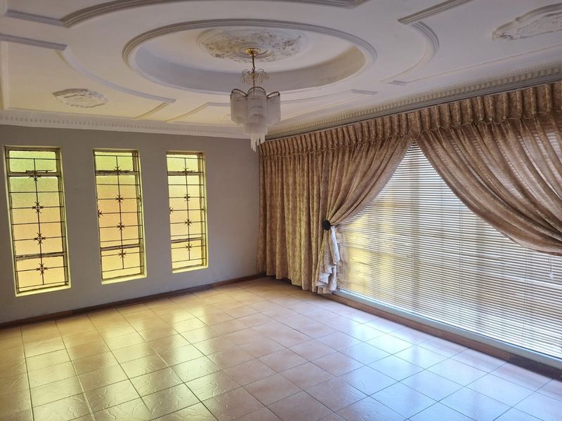 Spacious living room featuring ceramic tiled flooring, ornate ceiling detailing with a central chandelier, and large windows fitted with security bars and curtains.