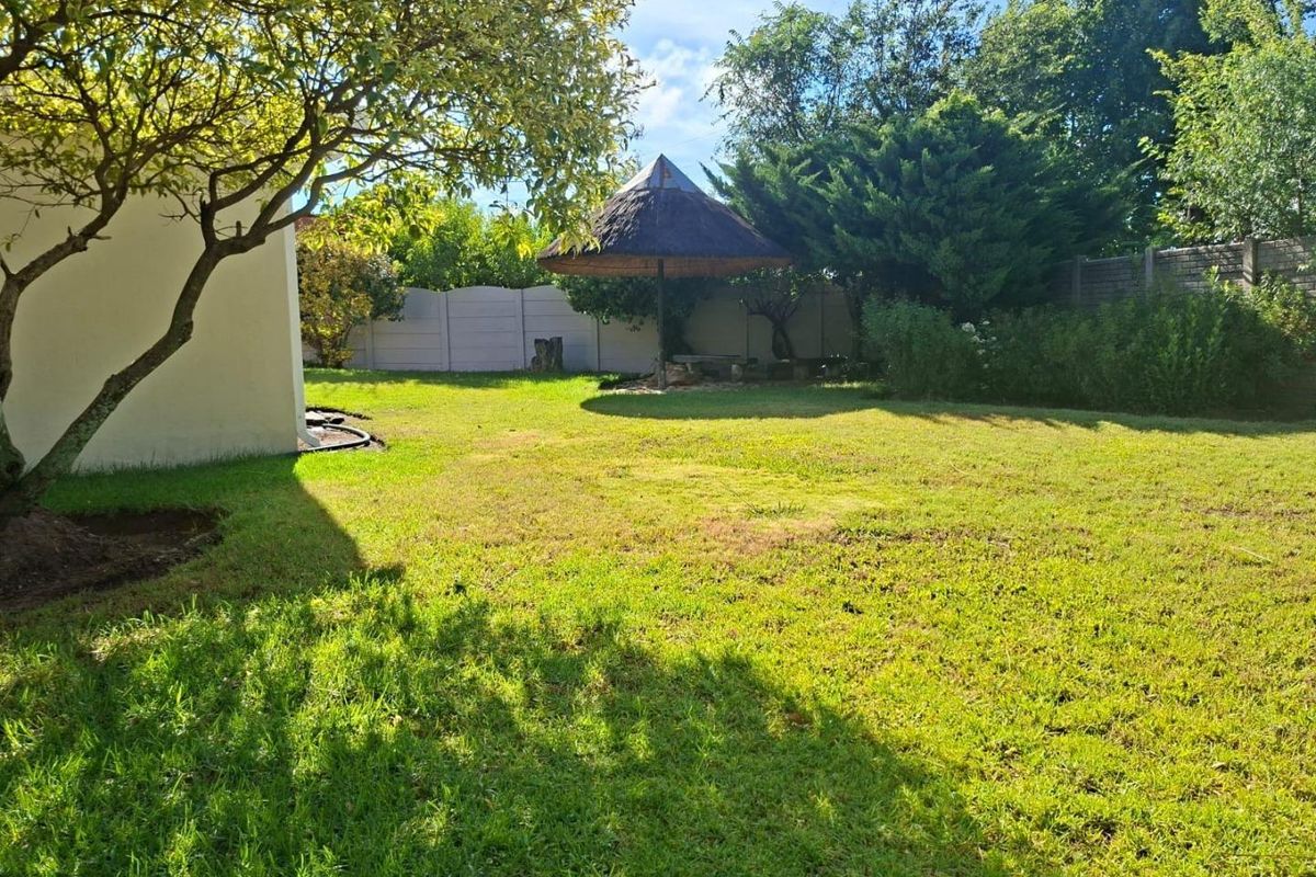A spacious residential garden featuring a well-maintained lawn, mature trees, and a traditional thatched lapa structure for outdoor relaxation.