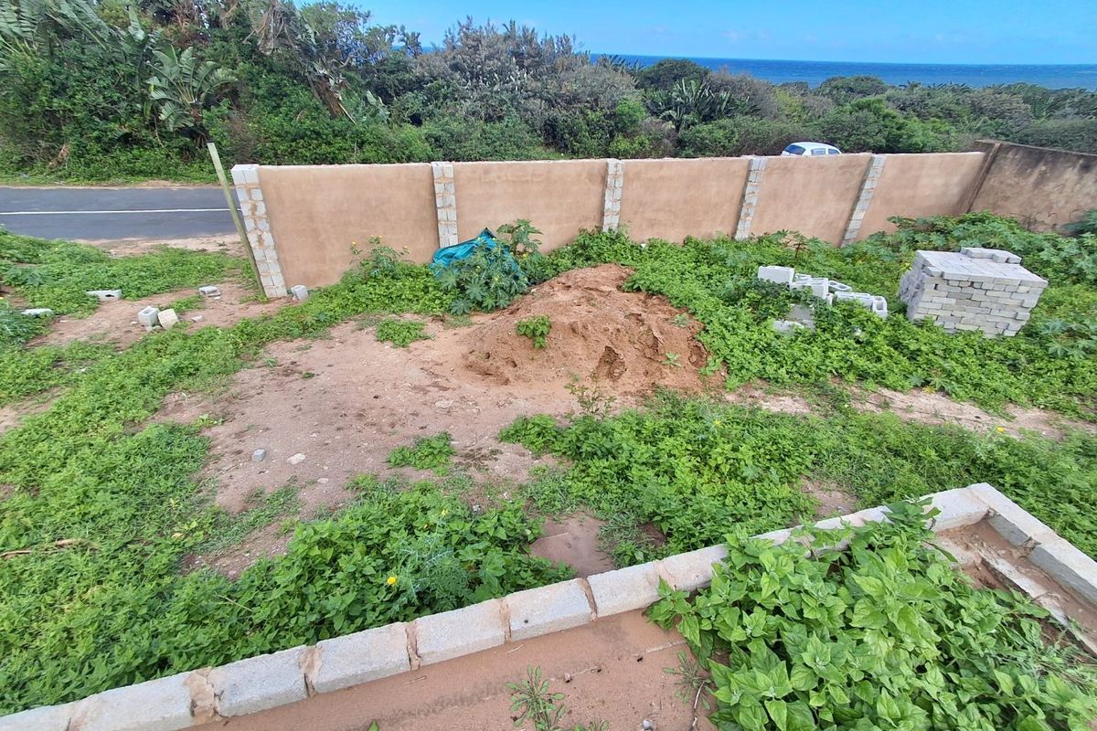 A view of a vacant residential plot featuring a perimeter boundary wall, overgrown vegetation, and an unimproved ground surface with construction materials.