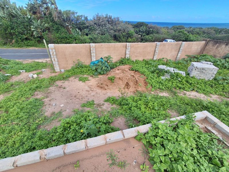 A view of a vacant residential plot featuring a perimeter boundary wall, overgrown vegetation, and an unimproved ground surface with construction materials.