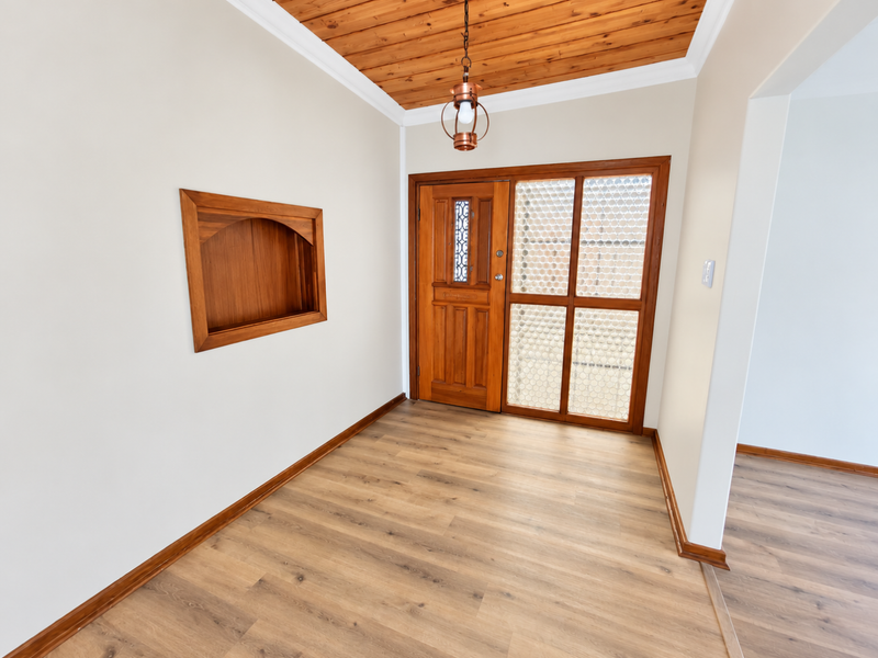 A bright and welcoming entrance hall featuring warm timber ceiling paneling, a classic wooden front door, and modern laminate flooring with a built-in wall niche.
