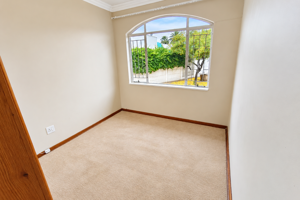A bright, empty bedroom featuring neutral walls, carpeted flooring, and an arched window with security bars overlooking the garden.