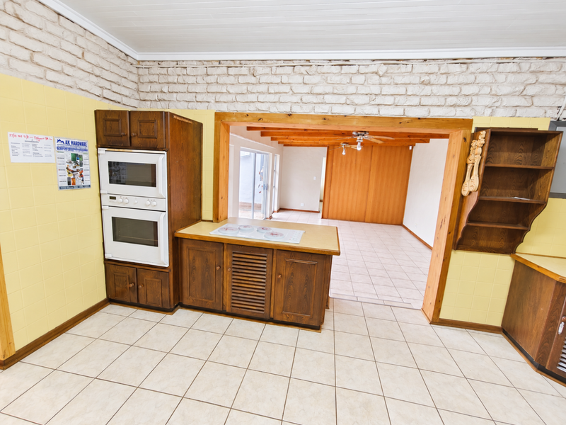 A dated kitchen featuring built-in double ovens, wooden cabinetry, and tiled surfaces, opening into an adjacent living area with exposed wooden ceiling beams.