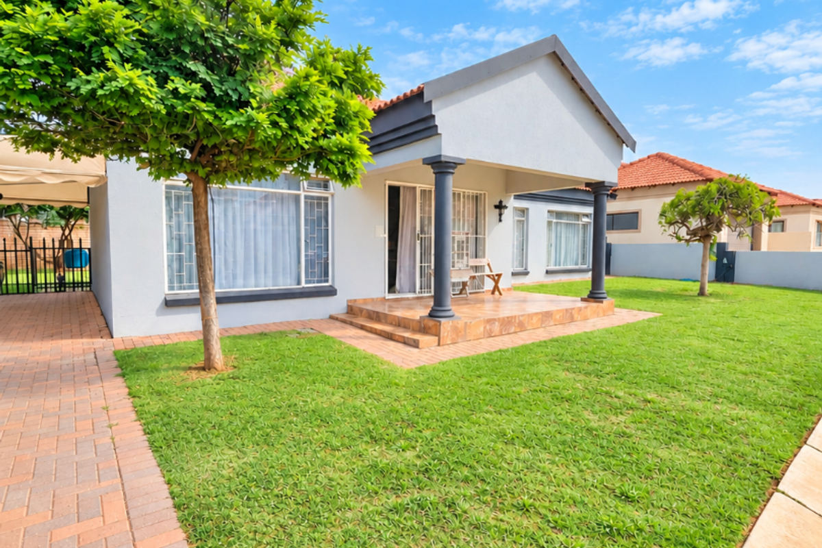 A well-maintained rear exterior view featuring a tiled patio, decorative columns, and a spacious lawn with paved driveway access.
