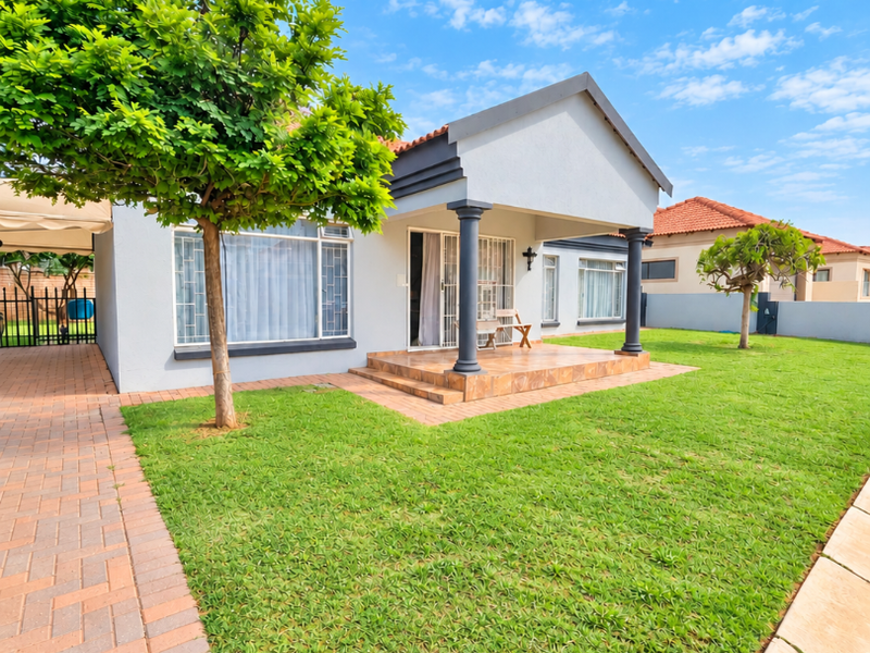 A well-maintained rear exterior view featuring a tiled patio, decorative columns, and a spacious lawn with paved driveway access.