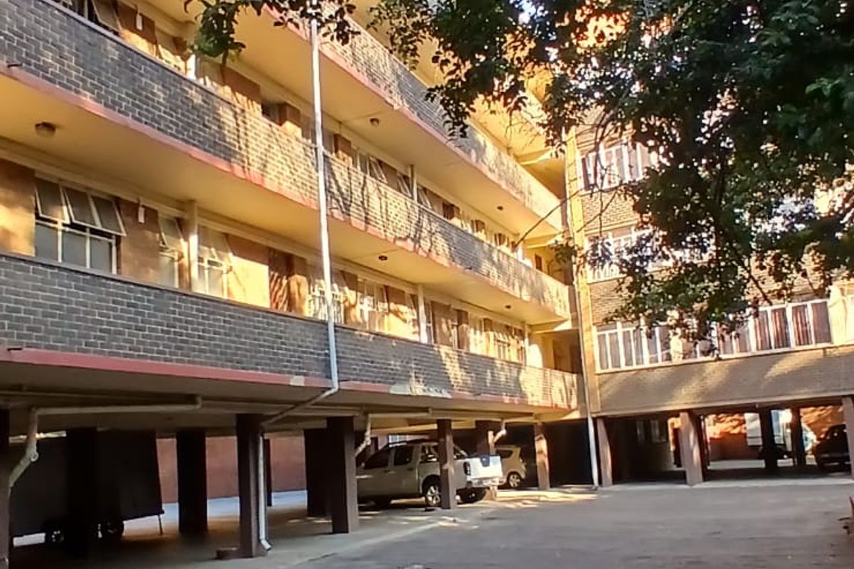 Exterior view of an apartment building featuring a brick facade, covered parking area, and significant wear to the asphalt ground surface.