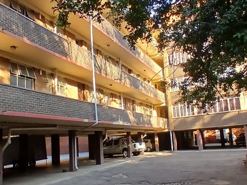 Exterior view of an apartment building featuring a brick facade, covered parking area, and significant wear to the asphalt ground surface.