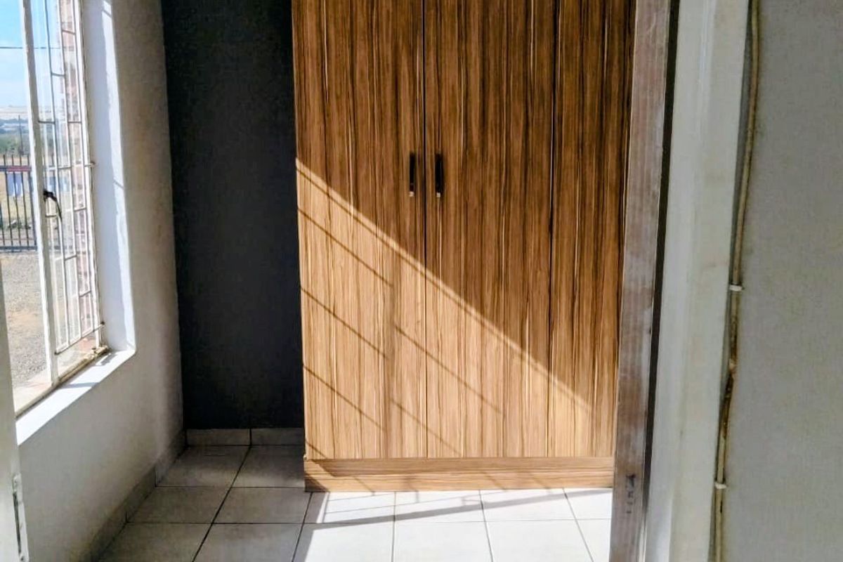 A compact bedroom featuring tiled flooring, a built-in wooden wardrobe, and natural light through a window with security bars.