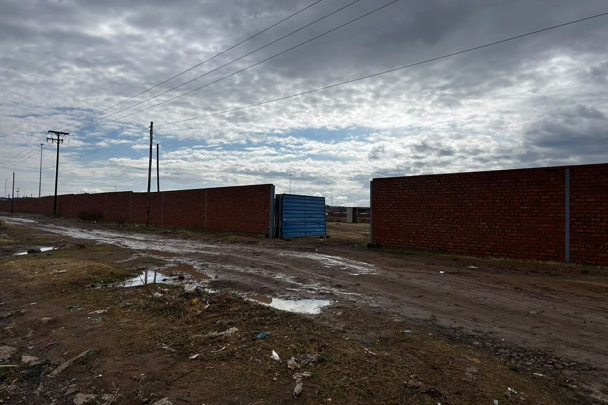 A view of an unpaved road leading to a property entrance enclosed by a brick boundary wall and metal gate.