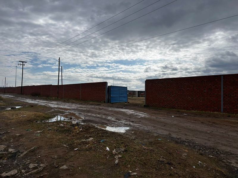 A view of an unpaved road leading to a property entrance enclosed by a brick boundary wall and metal gate.