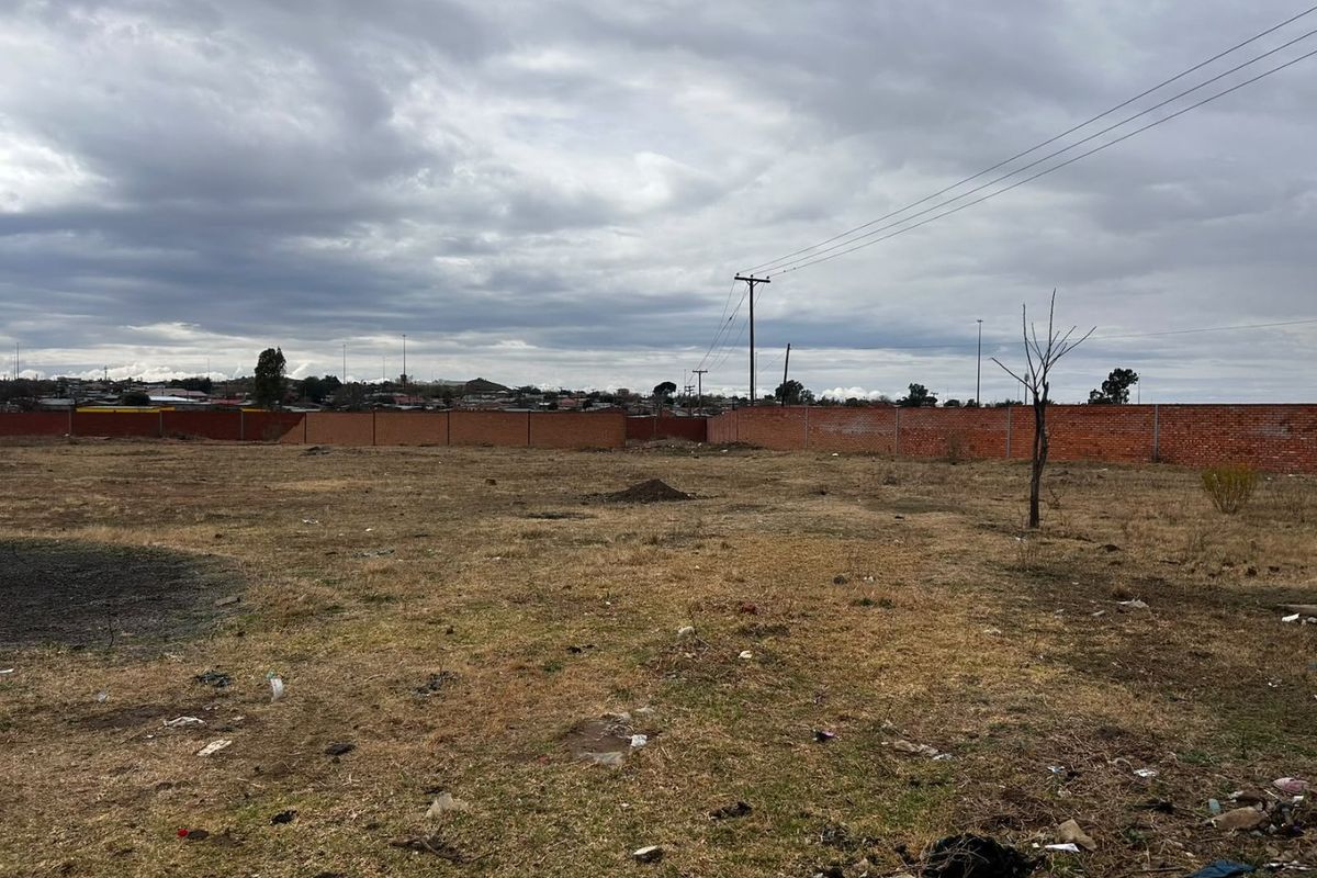 A large, vacant plot of land enclosed by a brick perimeter wall, featuring open ground and a utility pole.