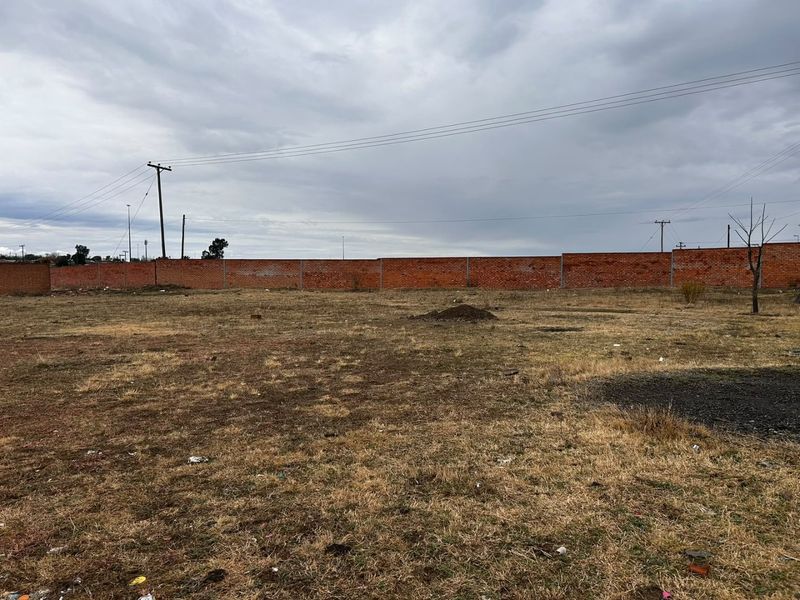 A large, undeveloped plot of land enclosed by a perimeter brick wall, featuring dry vegetation and utility infrastructure.