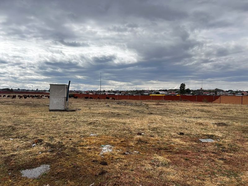 An expansive, undeveloped plot of land featuring a perimeter brick wall and a small utility kiosk, set against a suburban backdrop.