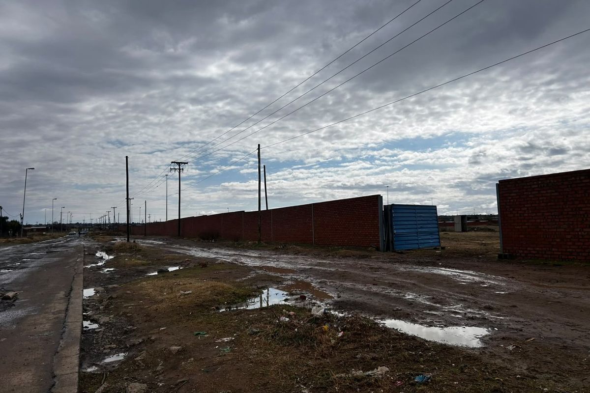 A street-level view showing an unpaved road, overhead power lines, and a long brick boundary wall under a cloudy sky.