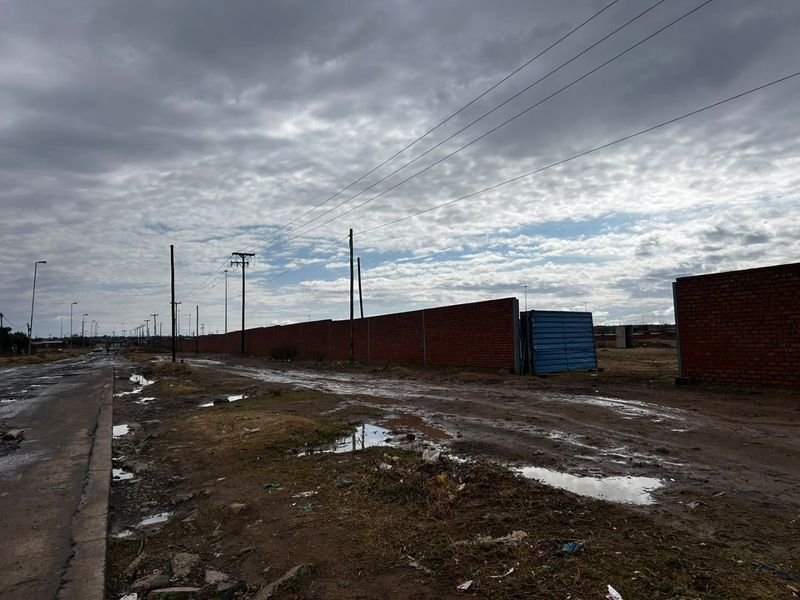 A street-level view showing an unpaved road, overhead power lines, and a long brick boundary wall under a cloudy sky.