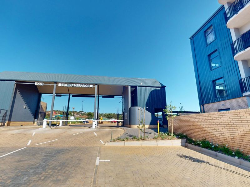 A modern apartment complex entrance featuring a secure gatehouse, paved driveway, and contemporary metal-clad building facade with balconies.