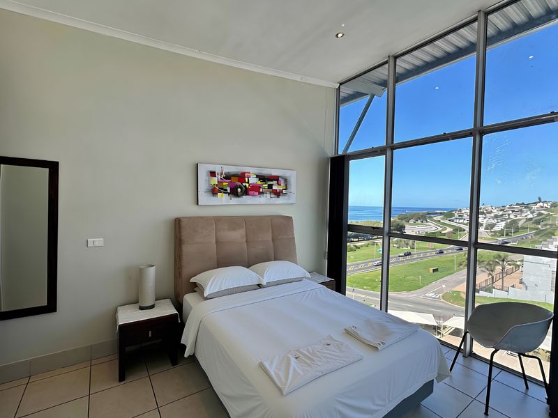 A bright bedroom featuring floor-to-ceiling windows with expansive coastal and road views, tiled flooring, and modern recessed lighting.