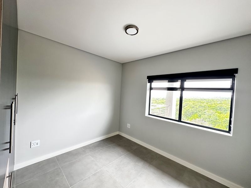 A clean, modern, and empty bedroom featuring neutral grey walls, large format floor tiles, a built-in cupboard, and a window with a roller blind.