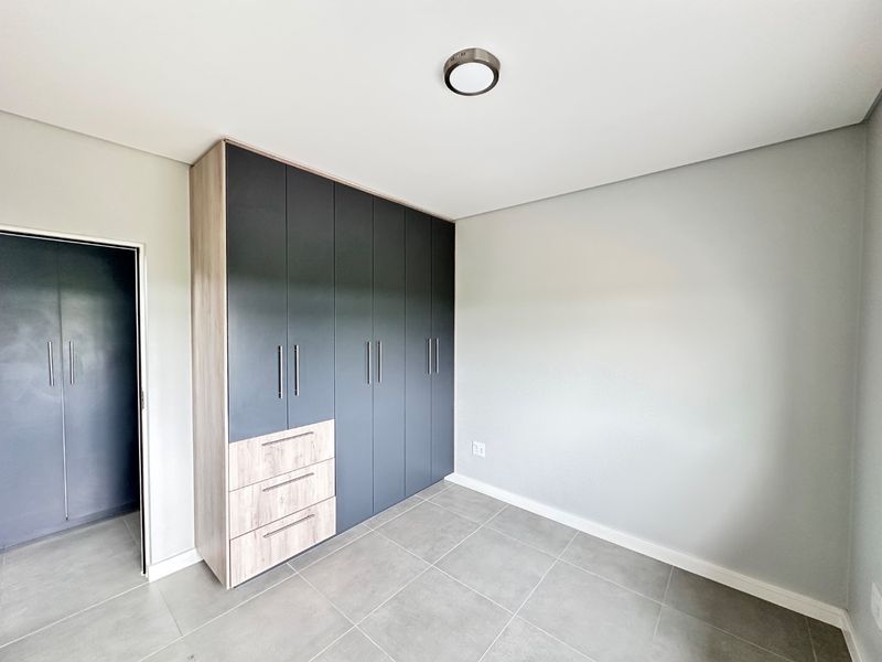 A clean, modern bedroom featuring neutral grey tiled flooring, light grey walls, and extensive dark grey built-in cabinetry with integrated drawers.