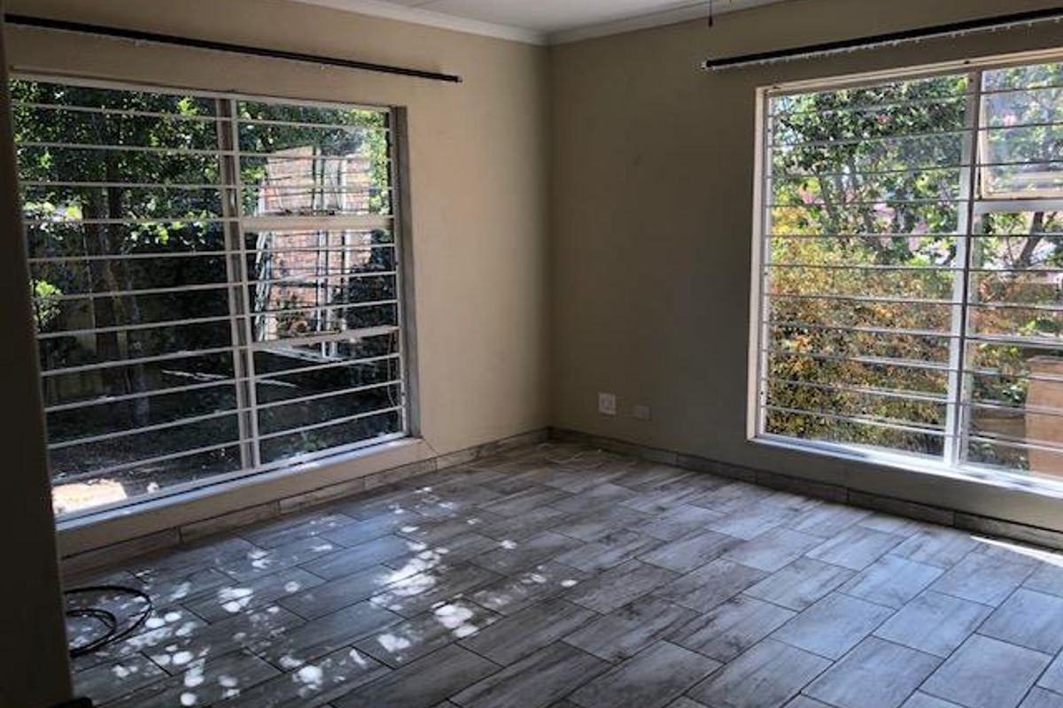 A bright, empty living room featuring modern wood-look tiled flooring, large windows with security bars, and neutral wall tones.