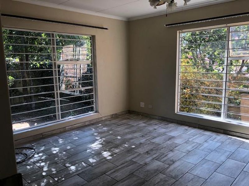 A bright, empty living room featuring modern wood-look tiled flooring, large windows with security bars, and neutral wall tones.