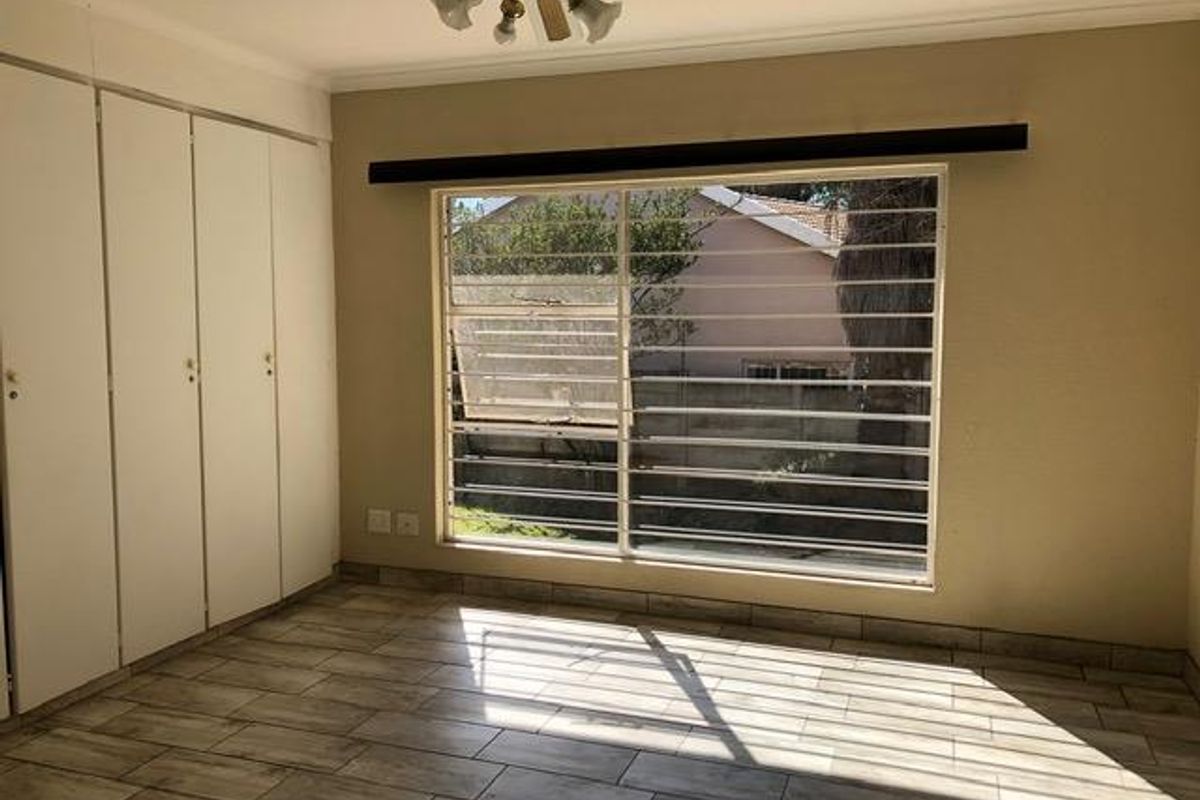 A bright, empty bedroom featuring tiled flooring, built-in cupboards, and a ceiling fan, with security bars installed on the window.