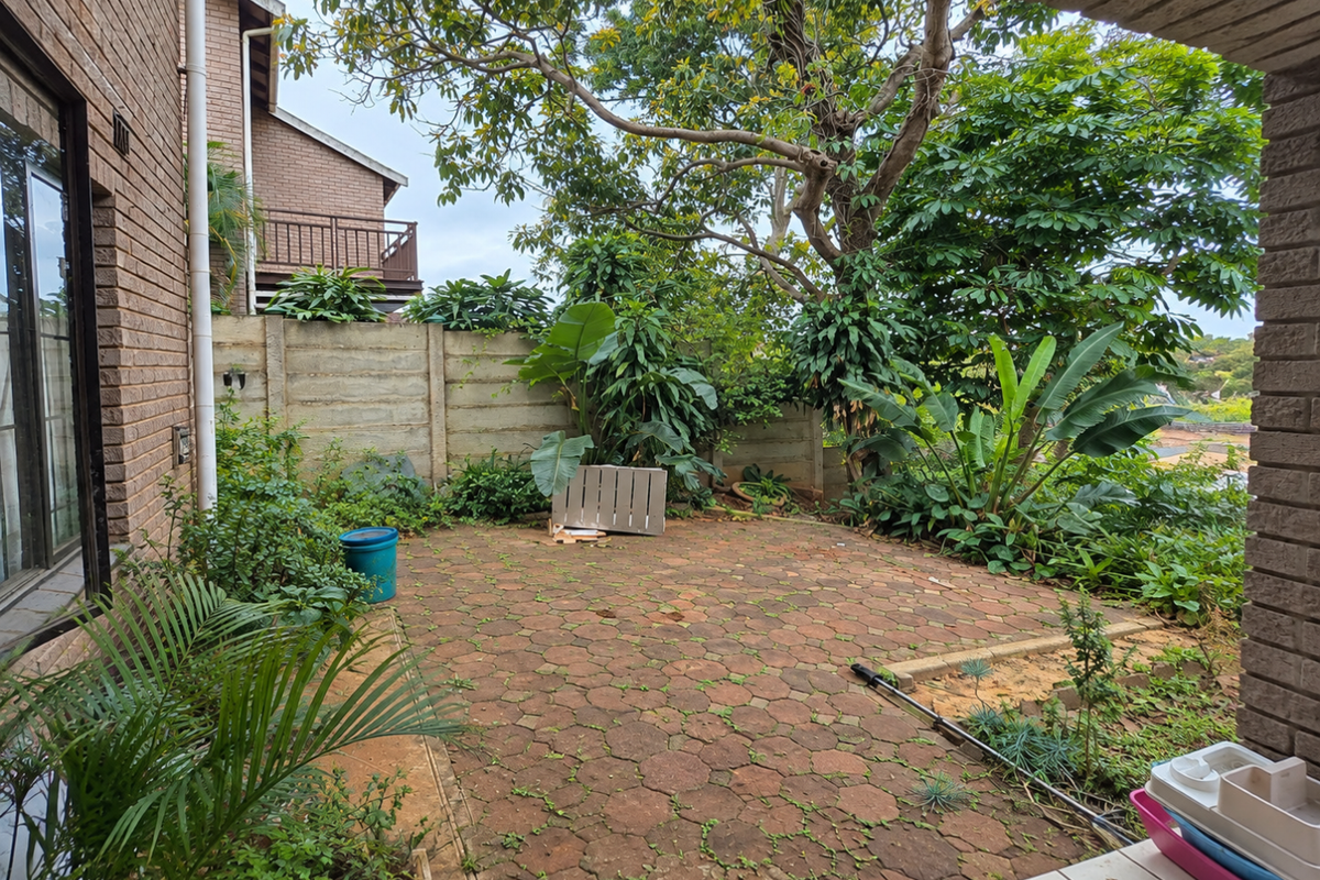 A private rear patio area featuring brick paving, established garden greenery, and a precast concrete boundary wall, offering potential for outdoor living.