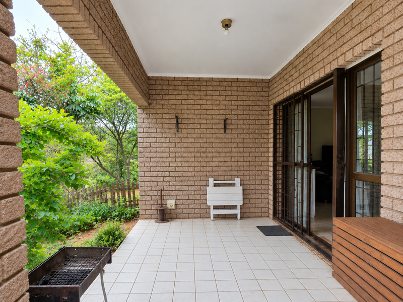 A covered patio featuring exposed brick walls, tiled flooring, and sliding glass door access to the interior, overlooking a private garden area.