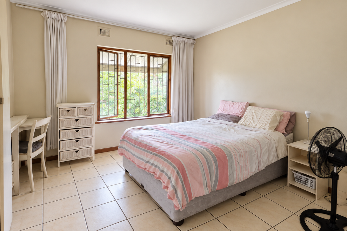 A well-lit bedroom featuring neutral wall tones, tiled flooring, and large windows with security bars, offering a functional and comfortable living space.