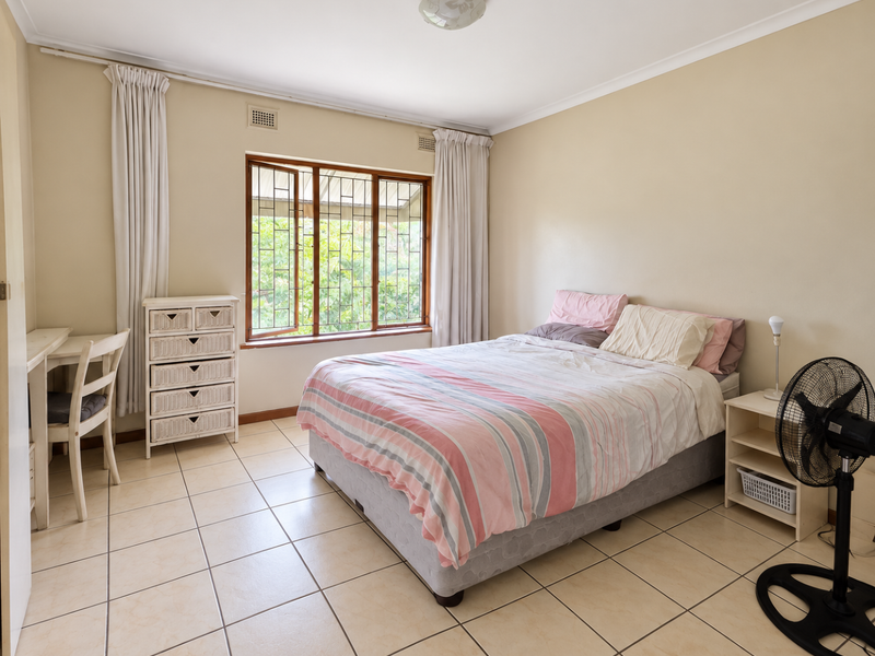 A well-lit bedroom featuring neutral wall tones, tiled flooring, and large windows with security bars, offering a functional and comfortable living space.