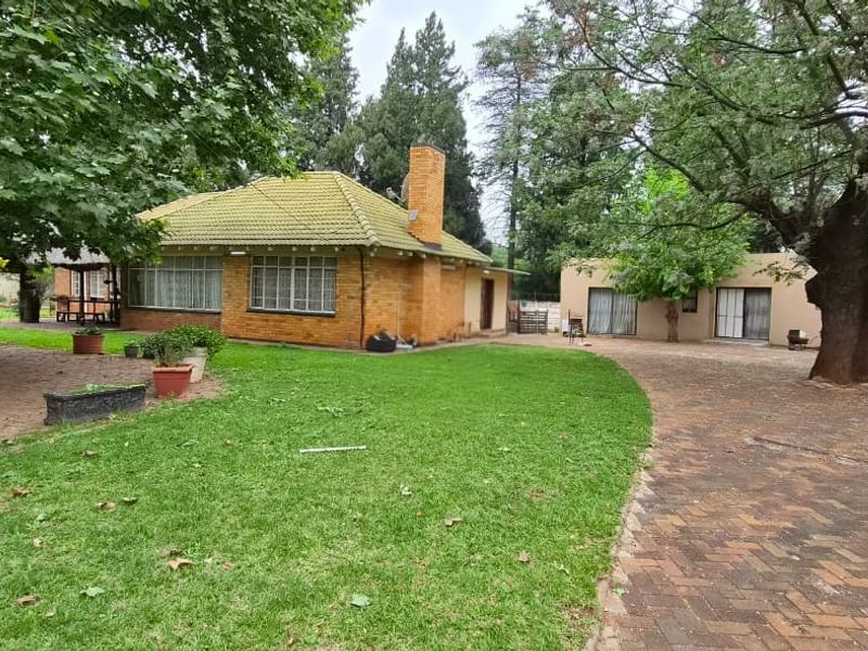 A view of the rear exterior featuring a brick house with a tiled roof, paved driveway, garage, and established garden with mature trees.