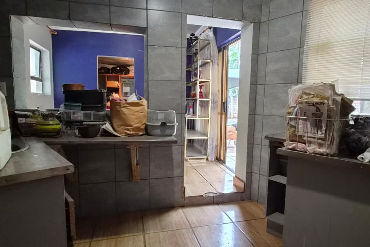 A dated kitchen area featuring grey wall tiles and tiled flooring, with a pass-through window and direct access to an adjacent room.