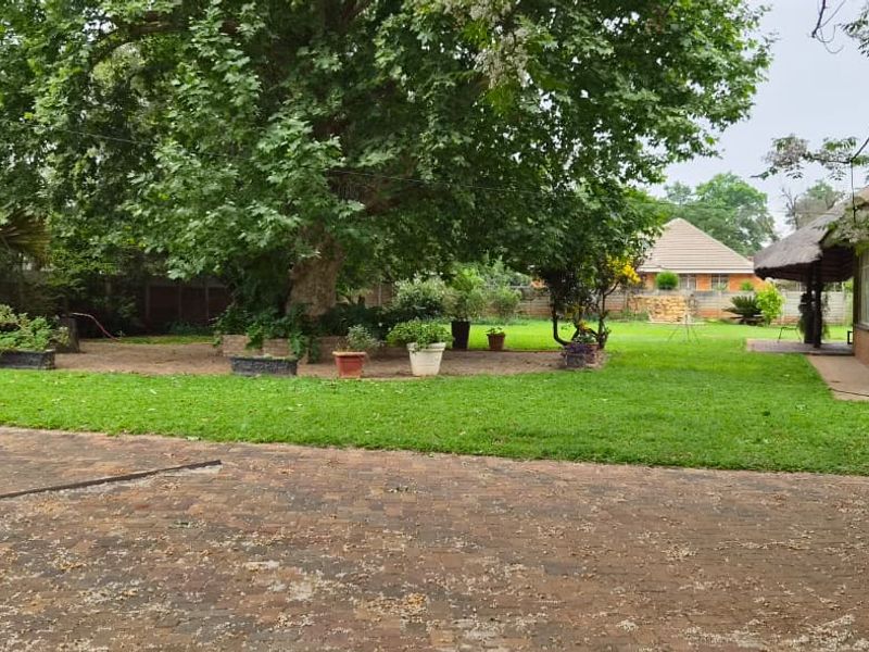 A spacious backyard featuring a large lawn, mature trees, and brick paving, adjacent to the main house structure.